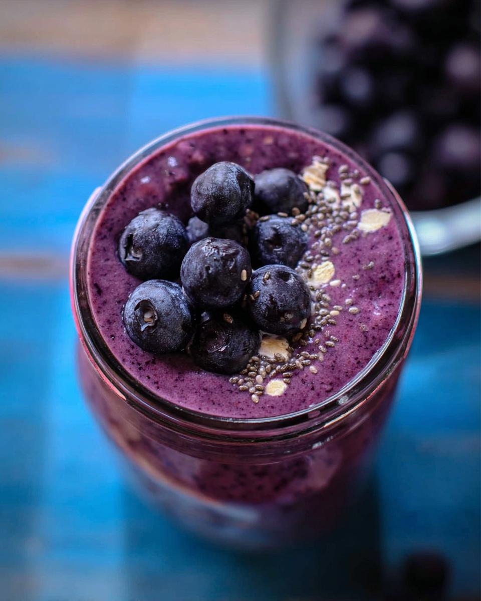 Overhead view of a thick, purple Blueberry Oat Breakfast Smoothie topped with fresh blueberries, chia seeds, and rolled oats.