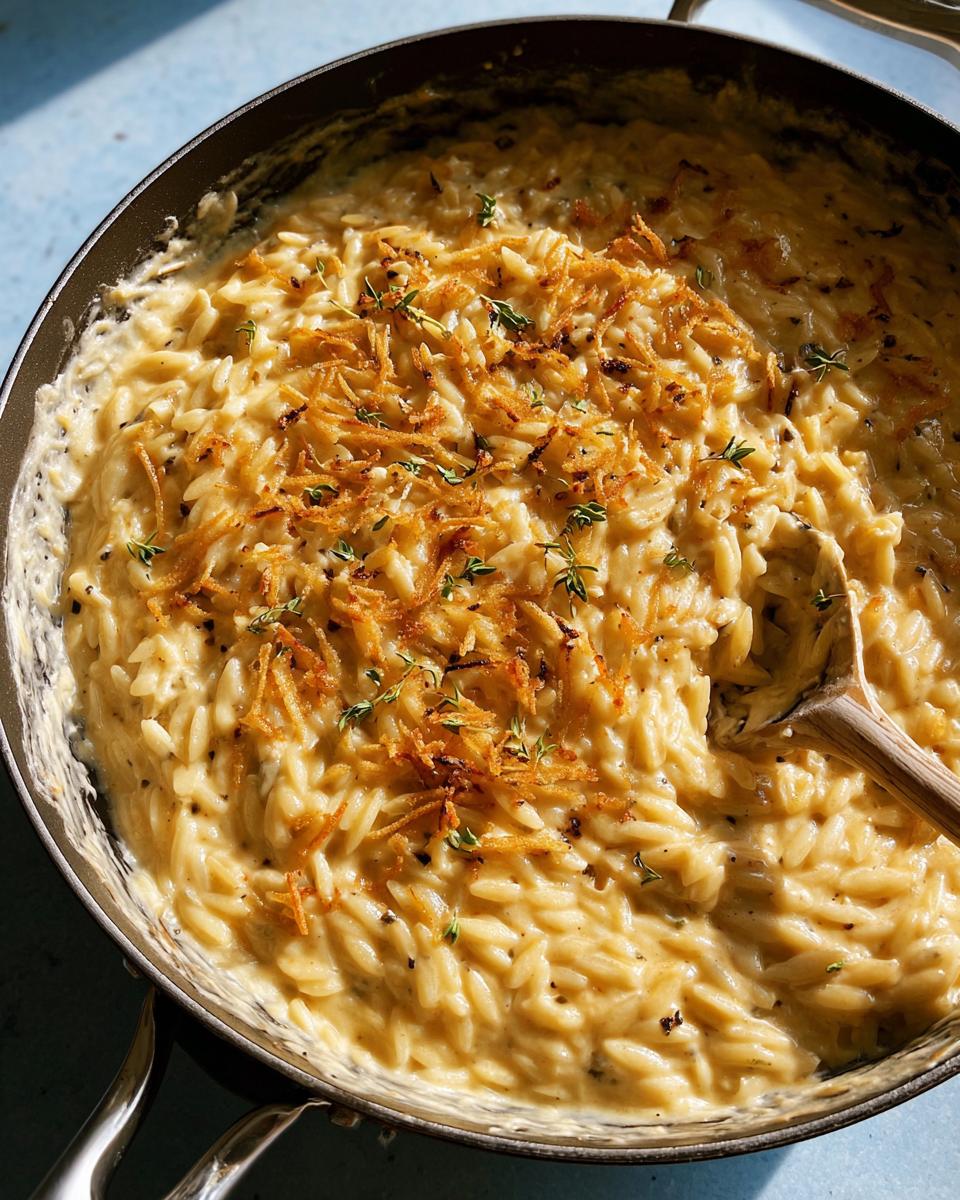 A close-up of creamy orzo pasta dish in a pan, topped with crispy fried onions and fresh thyme.