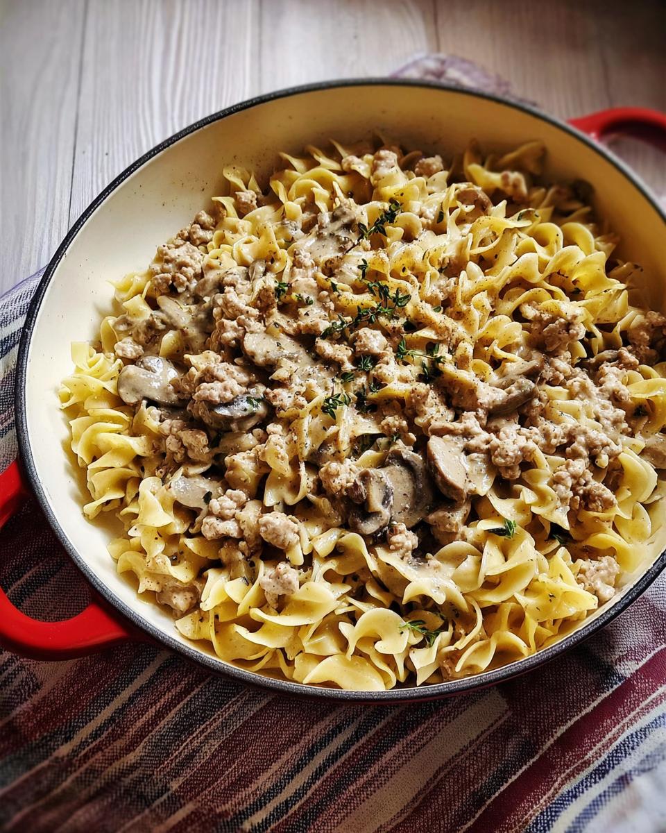 A close-up of Creamy Turkey Stroganoff with mushrooms and egg noodles in a red-handled pan, garnished with thyme.