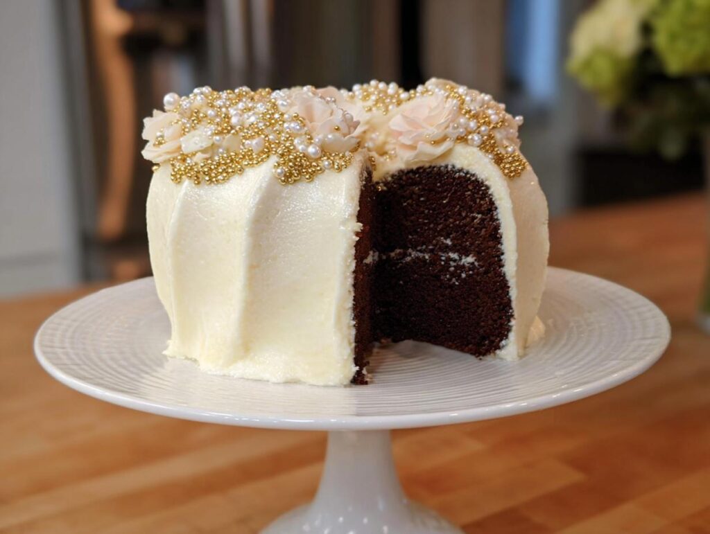 A rich, dark chocolate Gold-Dusted Bundt Cake with white frosting, decorated with gold sprinkles and blush flowers.