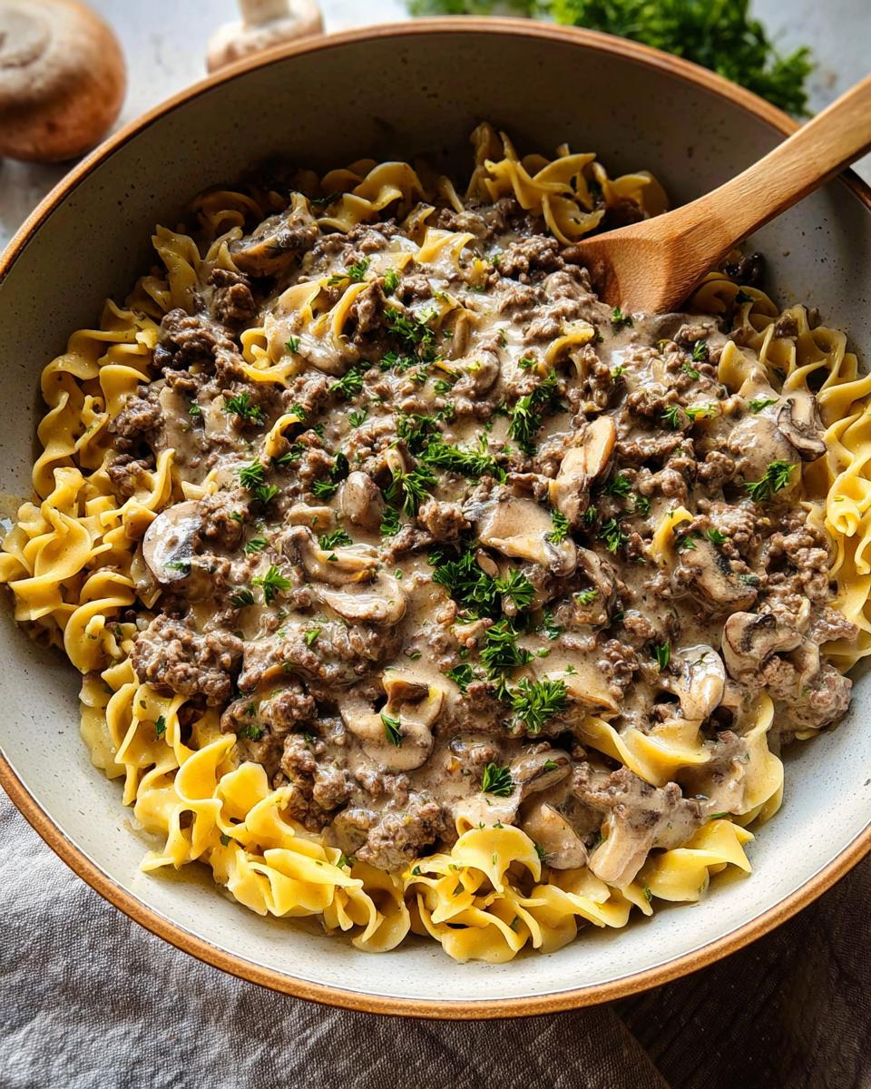 A close-up of a bowl filled with Ground Beef Stroganoff served over wide egg noodles, garnished with parsley.