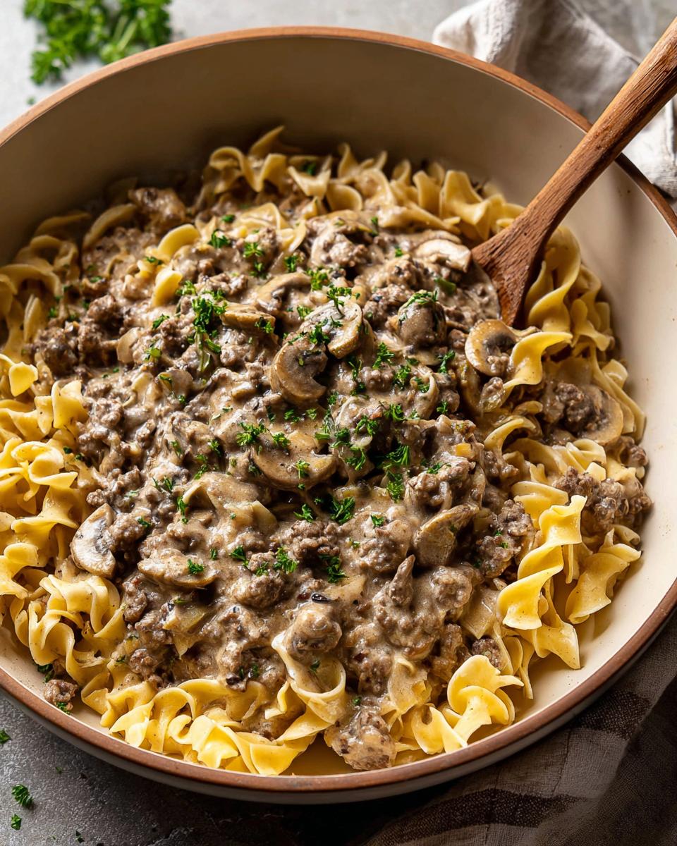 A close-up of rich Ground Beef Stroganoff served over wide egg noodles in a beige bowl.