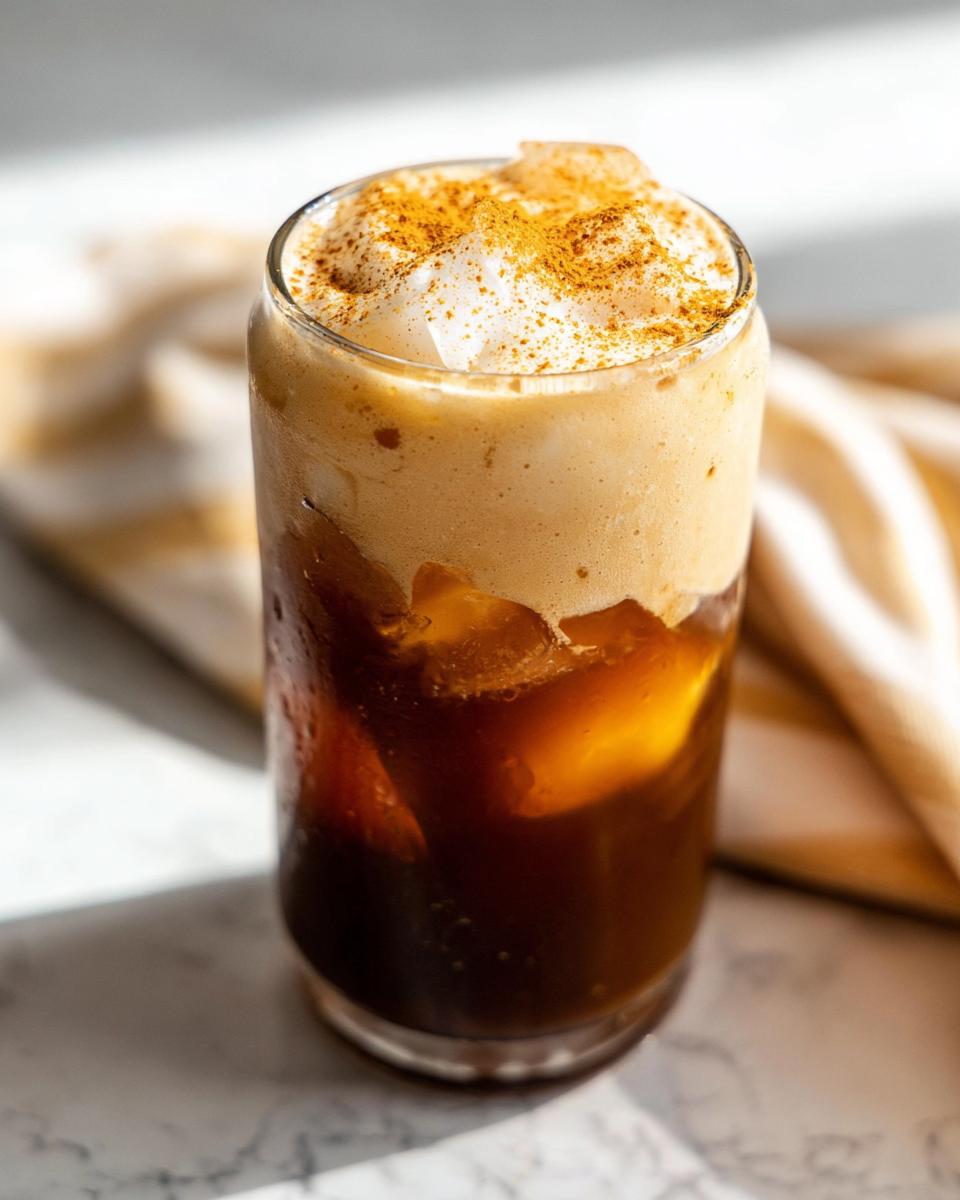 A tall glass of iced Pumpkin Cream Cold Brew topped with thick foam and sprinkled with cinnamon or pumpkin spice.