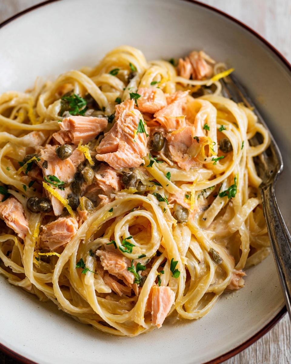 A close-up of Light & Creamy Salmon Pasta, featuring fettuccine noodles coated in a rich sauce, topped with flaked salmon, capers, and lemon zest.