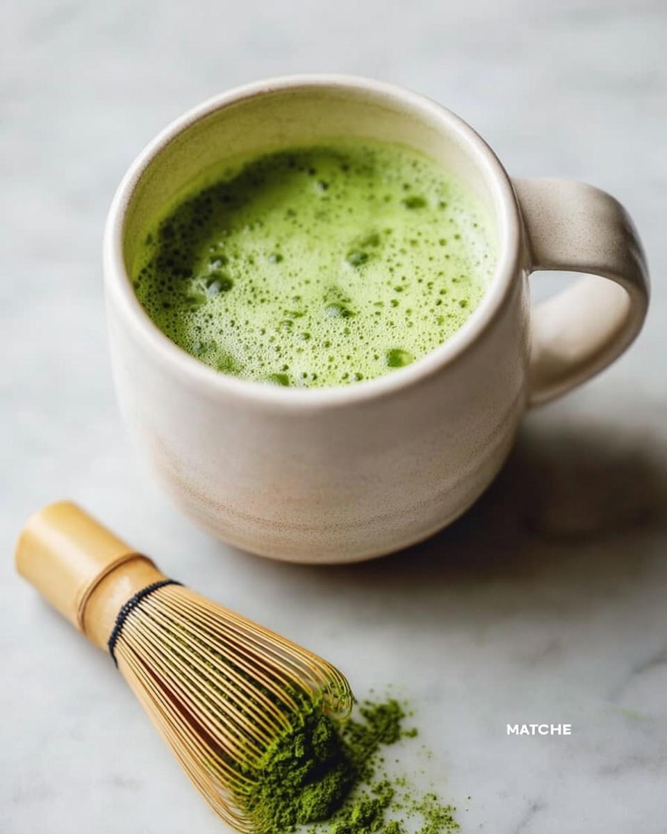 A finished Matcha Green Tea Latte in a ceramic mug next to a bamboo whisk with matcha powder.