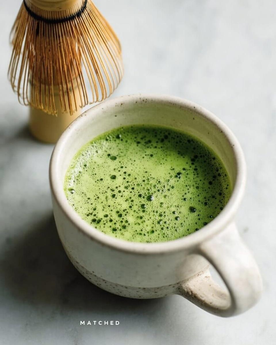 Close-up of a freshly prepared Matcha Green Tea Latte with vibrant green foam in a speckled ceramic mug, next to a bamboo whisk.