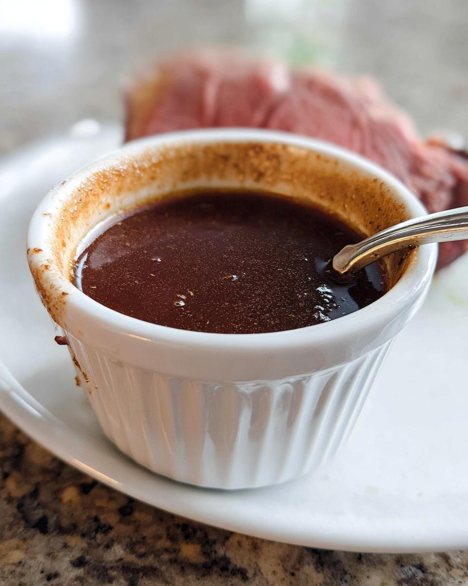 Close-up of rich, dark brown Prime Rib Au Jus Recipe served in a white fluted ramekin with a spoon.