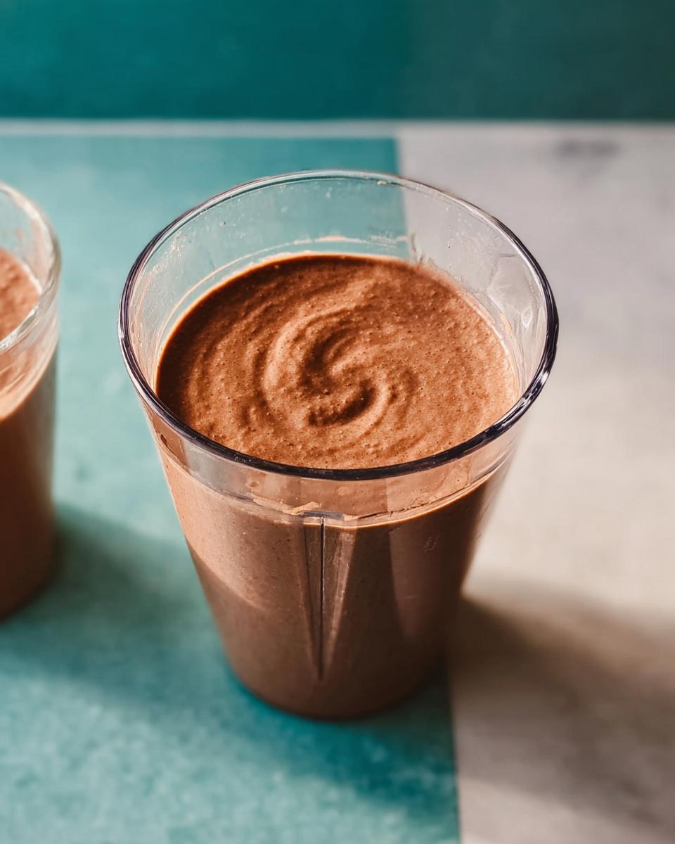 Close-up of a thick, brown Peanut Butter Protein Smoothie with a swirl pattern on top, in a clear glass.