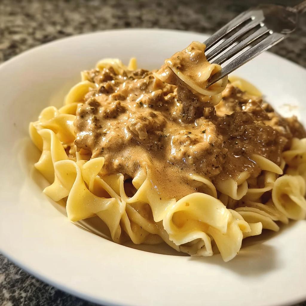 A fork lifts a bite of rich, creamy turkey stroganoff sauce and egg noodles from a white bowl.