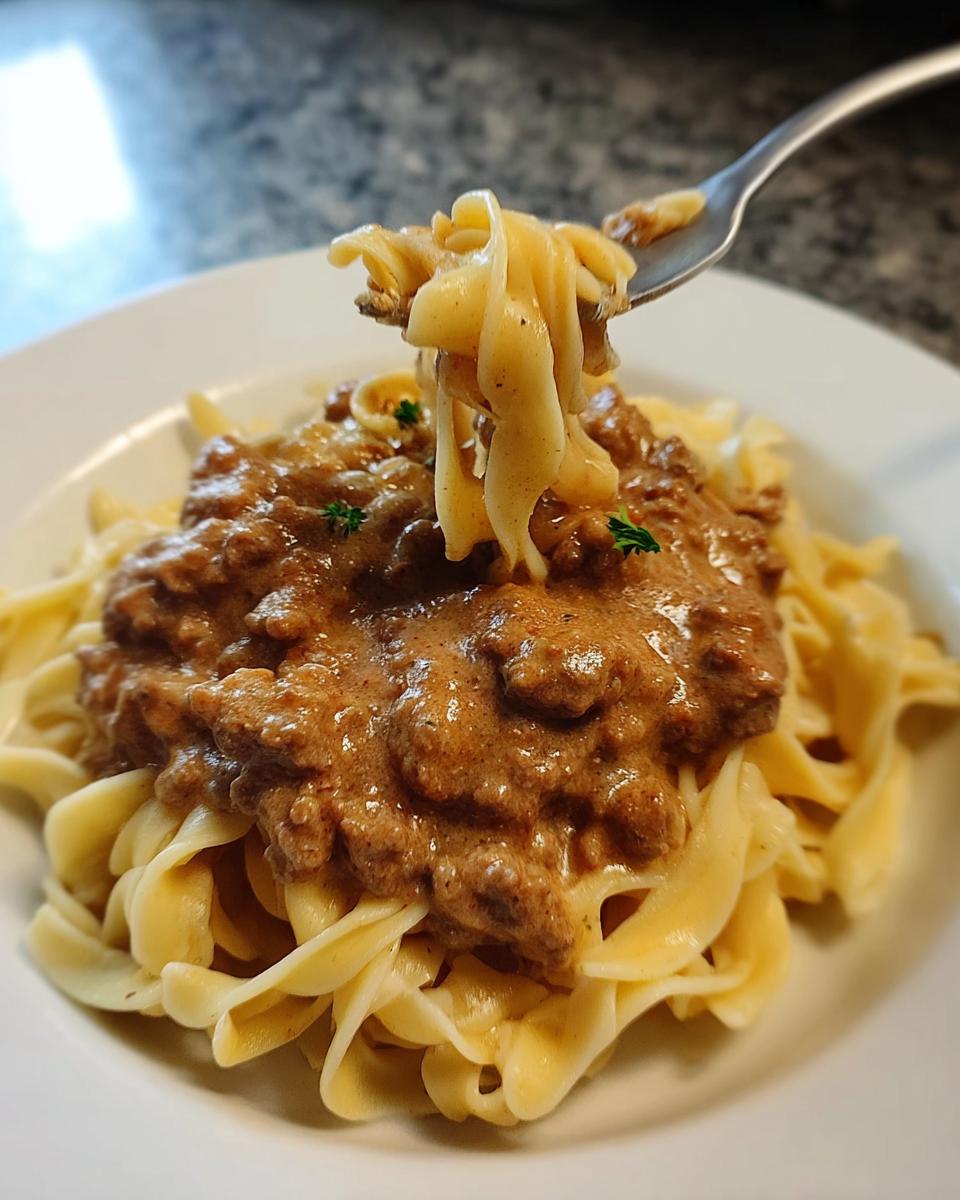 A fork lifts a bite of creamy turkey stroganoff and egg noodles from a white plate.