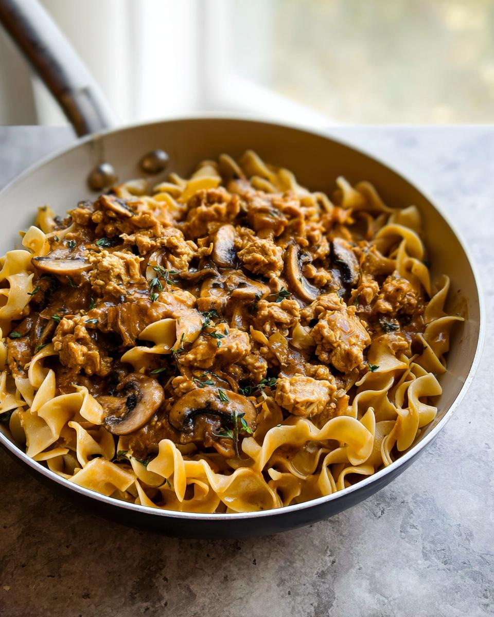 Close-up of Creamy Turkey Stroganoff served over wide egg noodles in a skillet, garnished with mushrooms and thyme.