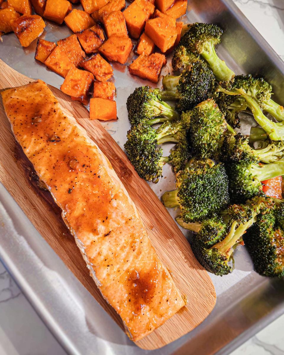 A cooked salmon fillet glazed with maple dijon sauce rests on a wooden plank next to roasted broccoli and sweet potatoes in a sheet pan dinner.
