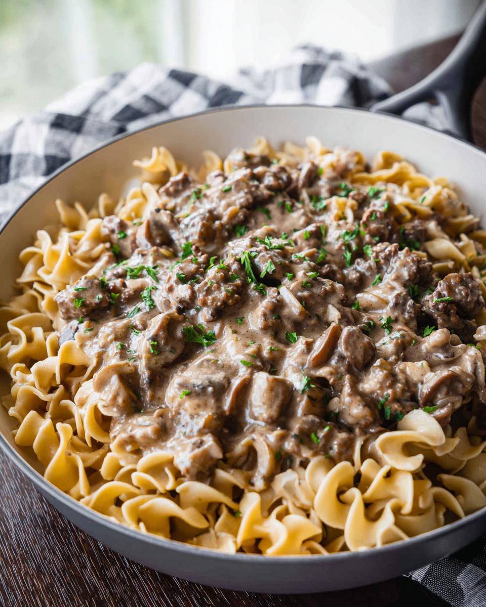 A close-up of One Pot Ground Beef Stroganoff served over wide egg noodles in a skillet, garnished with parsley.
