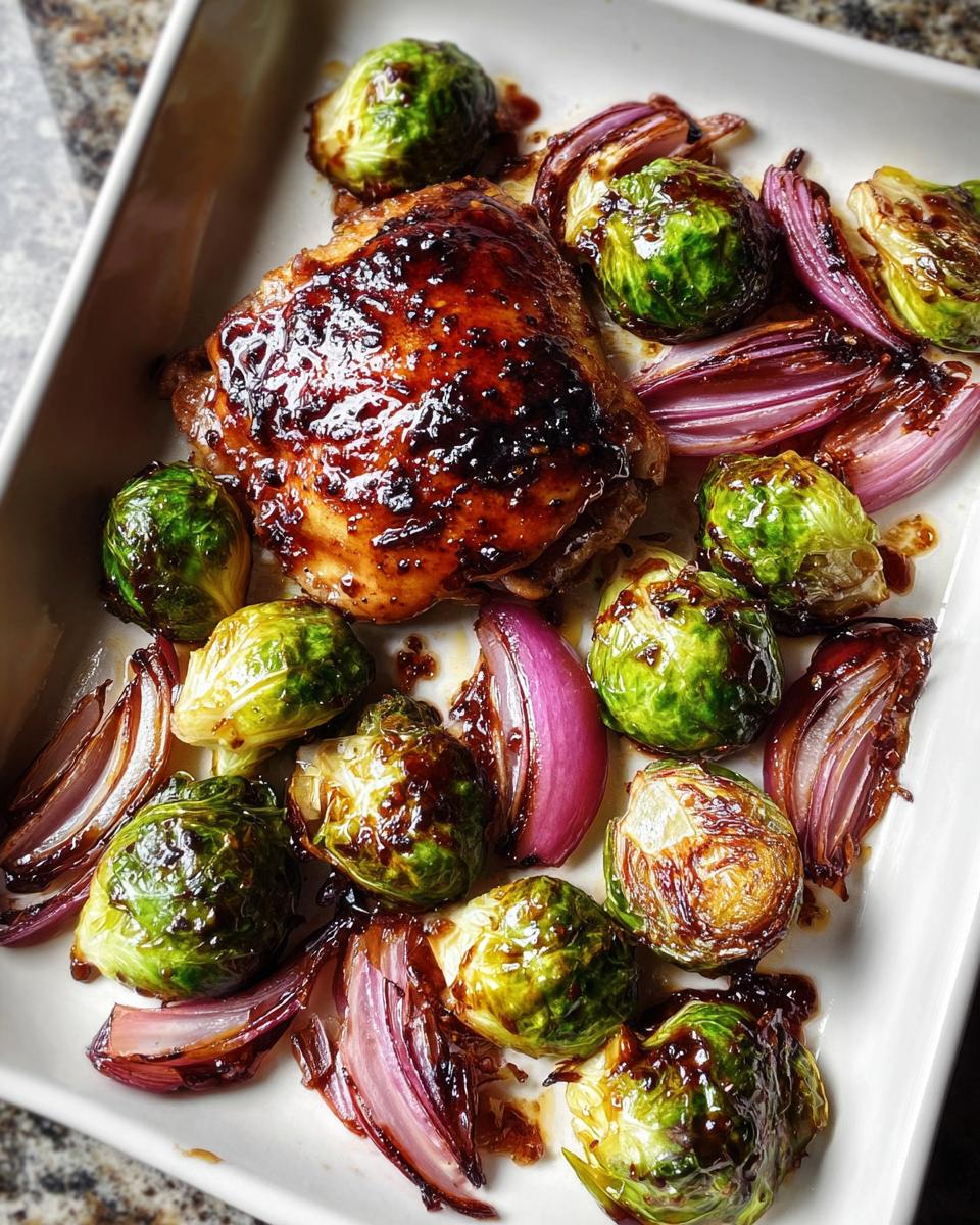 Close-up of glazed Sheet Pan Balsamic Chicken and Brussels Sprouts with red onion wedges in a white baking dish.