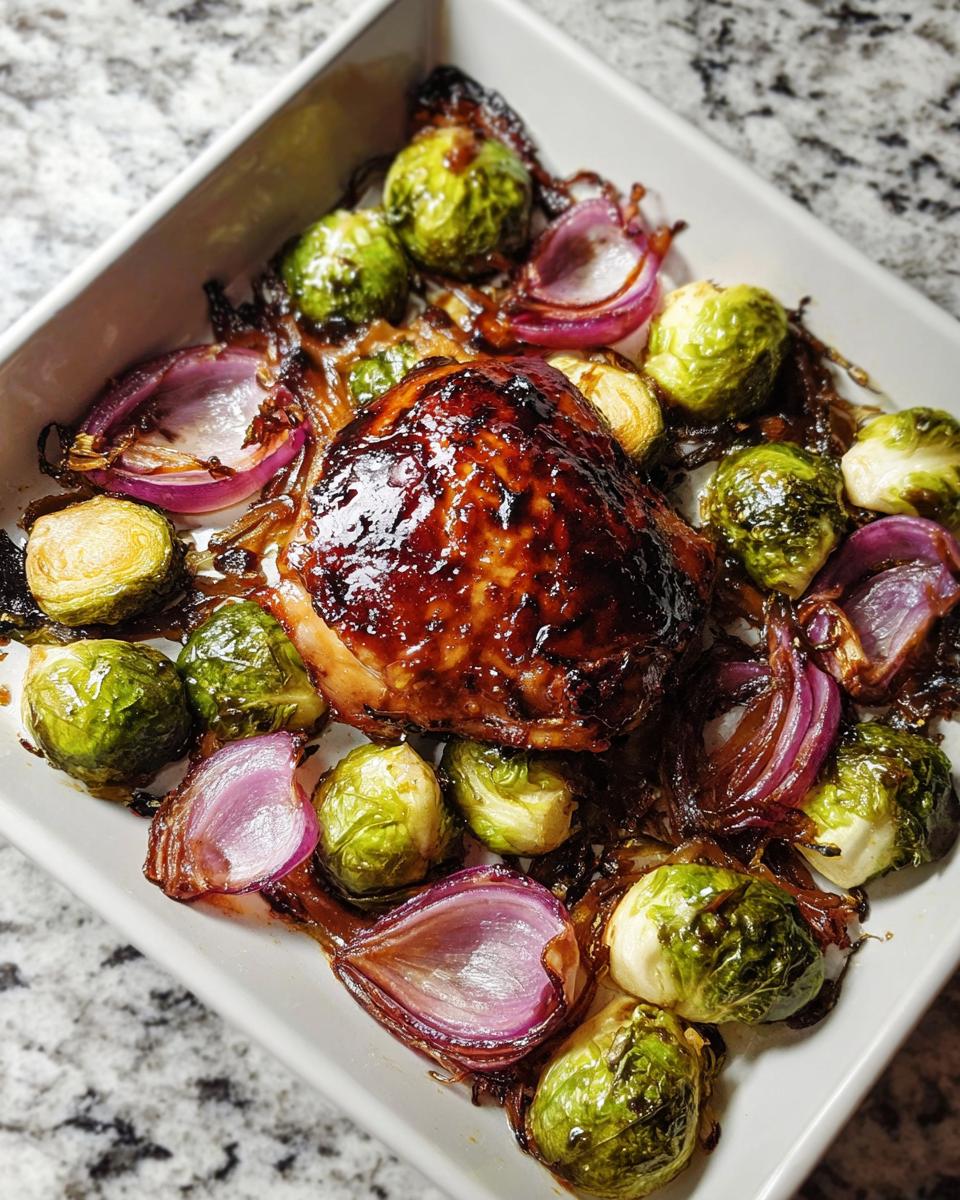 A close-up of a glazed chicken thigh surrounded by roasted Brussels sprouts and red onion wedges from Sheet Pan Balsamic Chicken and Brussels Sprouts.