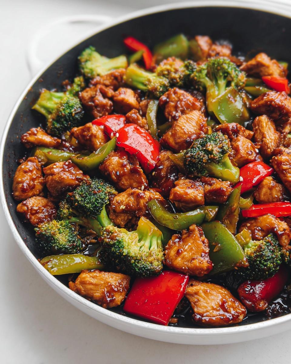 Close-up of chunks of chicken coated in teriyaki sauce mixed with bright broccoli florets and colorful red and green bell peppers in a skillet.