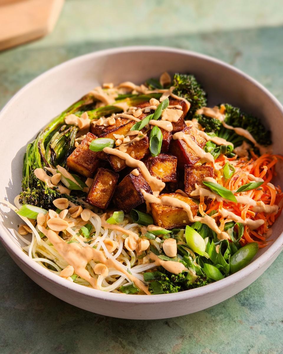 A vibrant bowl of 20-Ingredient Rice Bowls Recipe featuring crispy tofu, broccoli, shredded carrots, noodles, and a creamy peanut sauce.