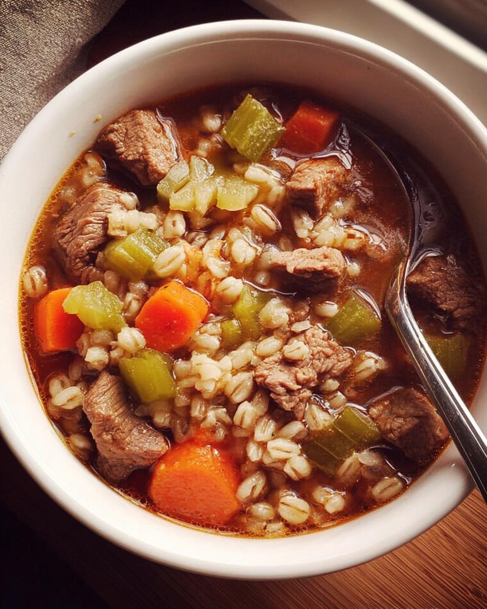 A close-up overhead view of a bowl of beef and barley soup, featuring tender chunks of beef, carrots, celery, and barley in a rich broth. Perfect for soup recipes meal prep.