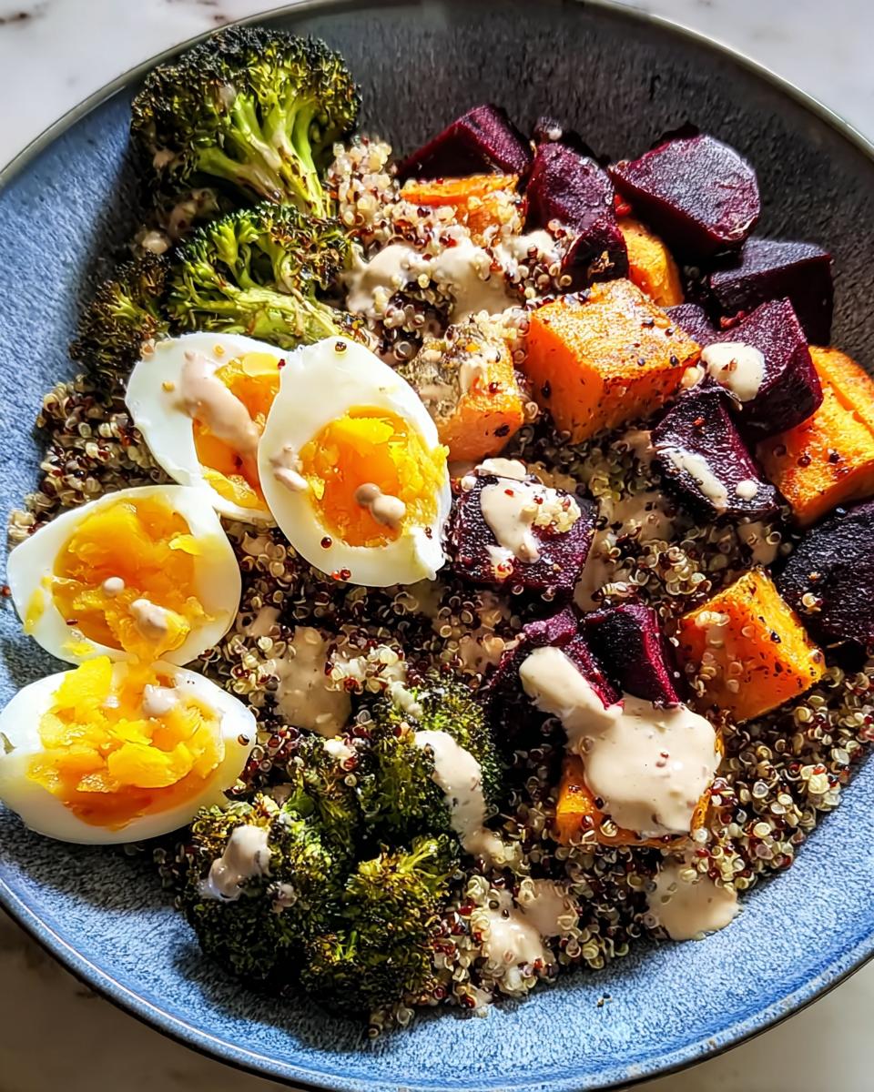 A vibrant bowl of breakfast ideas featuring quinoa, roasted broccoli, beets, sweet potato, and hard-boiled eggs drizzled with a creamy sauce.