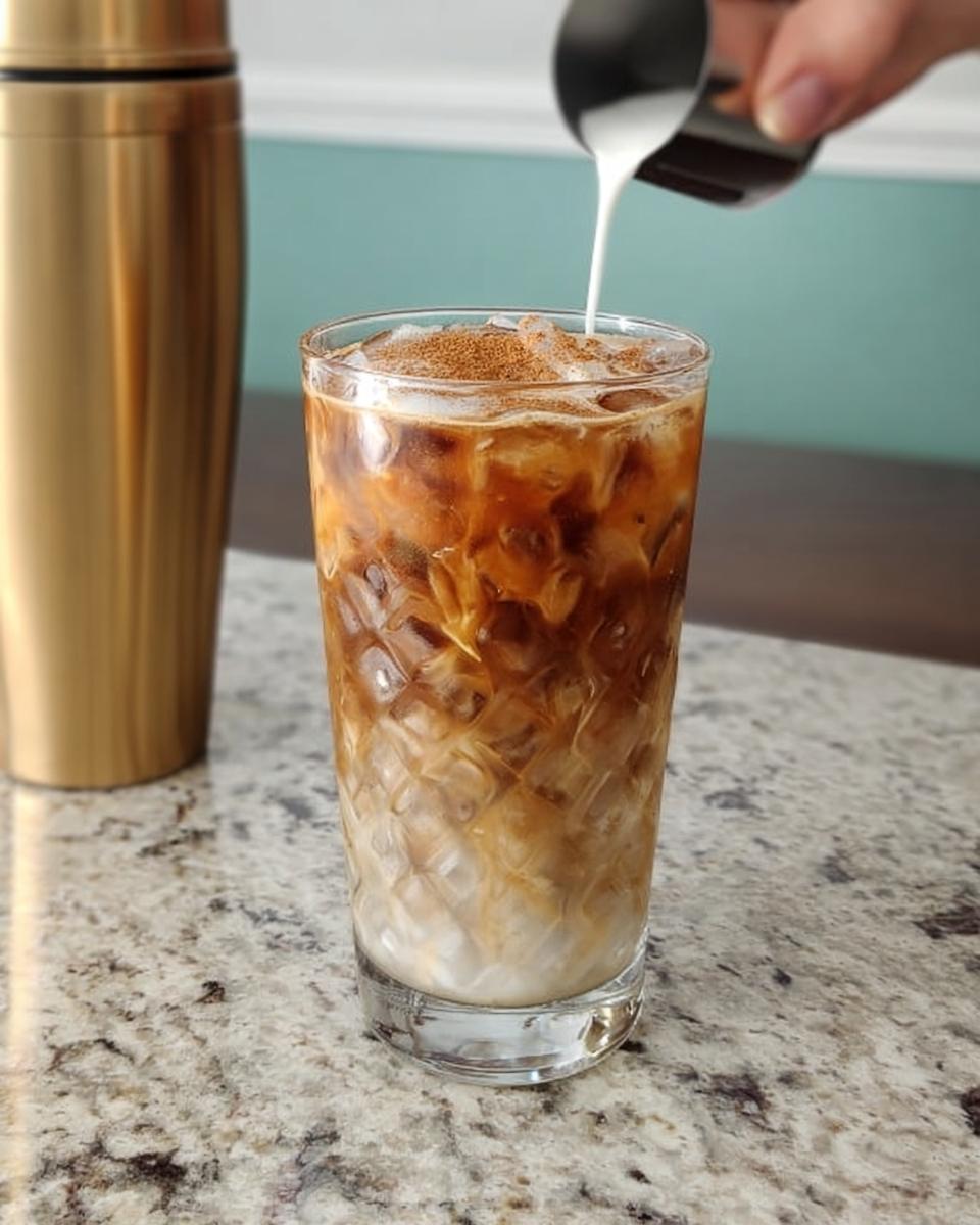 A refreshing iced coffee drink with layers of milk and coffee, topped with cinnamon, being prepared.