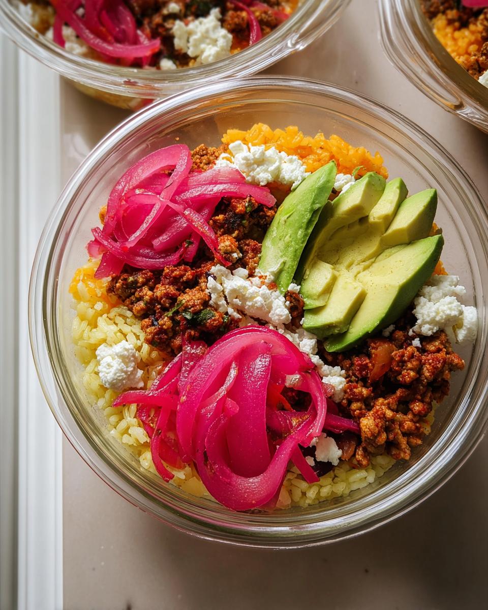 Overhead view of a meal prep rice bowl with yellow rice, seasoned ground meat, avocado slices, crumbled white cheese, and bright pink pickled onions.
