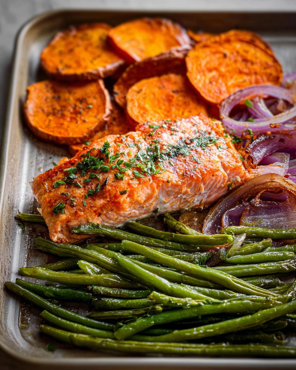 A close-up of a delicious salmon fillet meal prep with roasted sweet potatoes, green beans, and red onions on a baking sheet.
