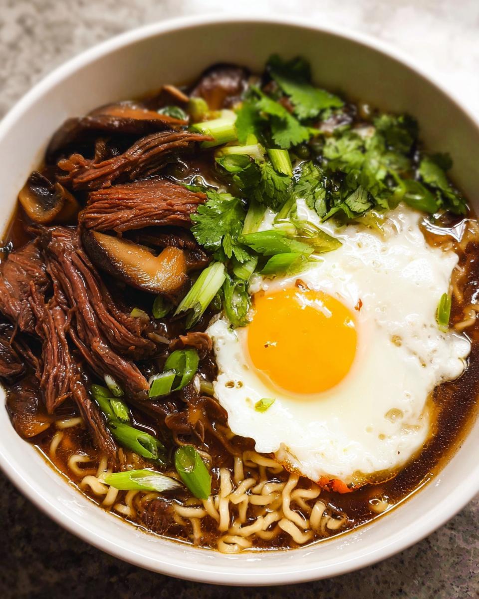 A close-up of a bowl of Slow Cooker Beef Ramen Noodles topped with shredded beef, mushrooms, green onions, cilantro, and a fried egg.