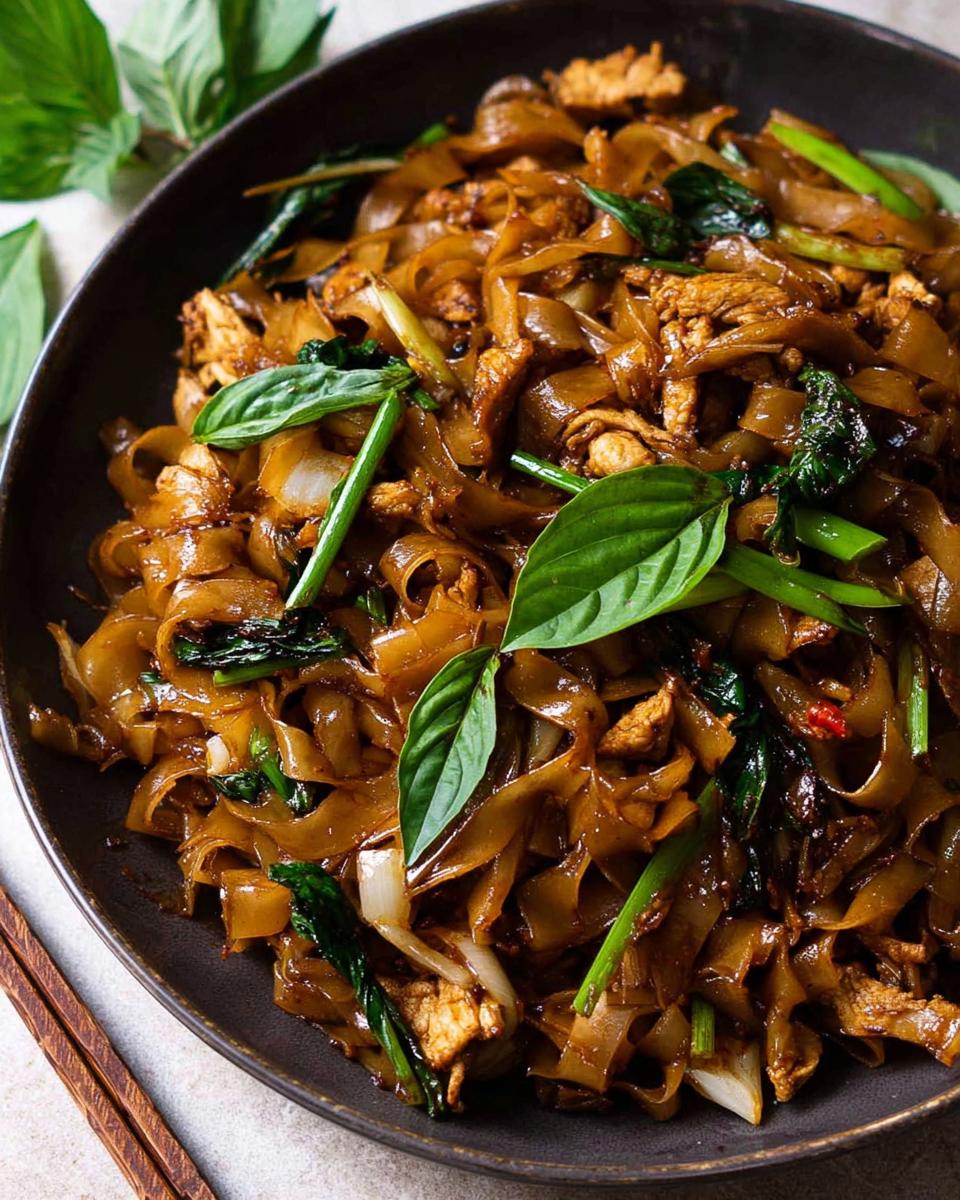 A close-up of a dark bowl filled with glossy, wide rice noodles, chicken, and fresh basil, representing Street Style Thai Drunken Noodles.