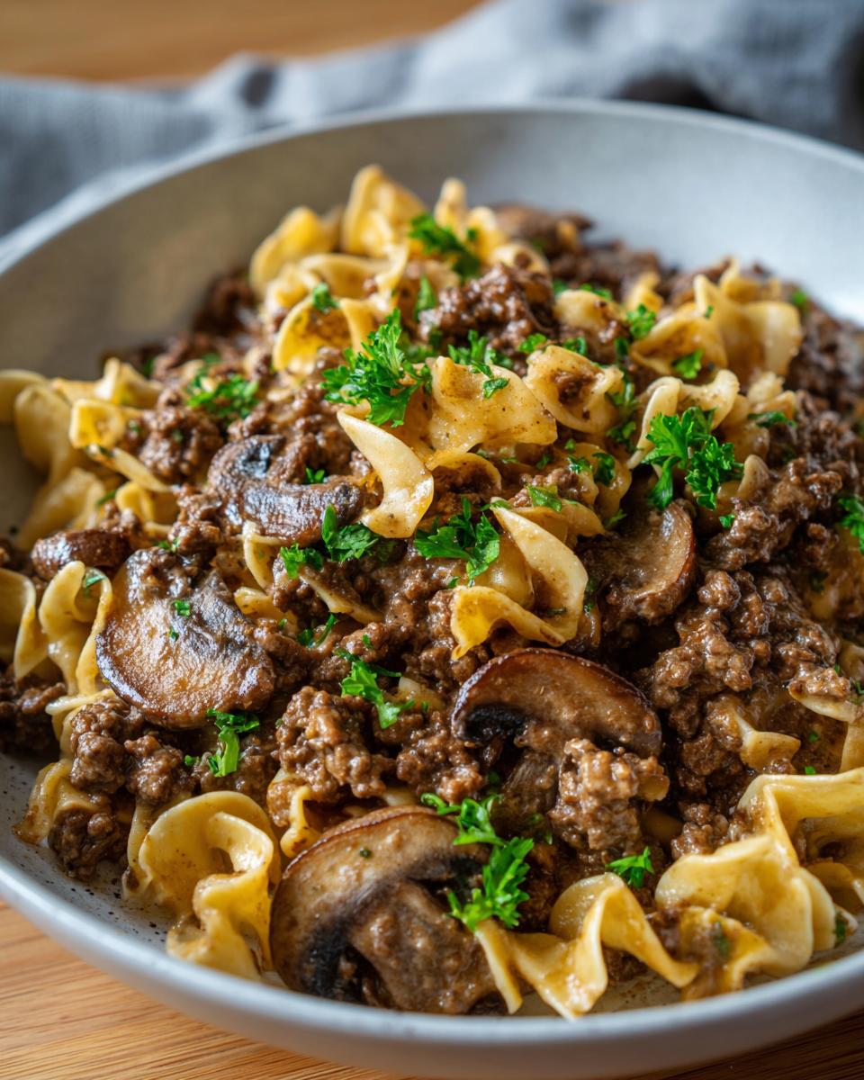 Close-up of a bowl filled with creamy beef stroganoff, featuring ground beef, egg noodles, mushrooms, and parsley.