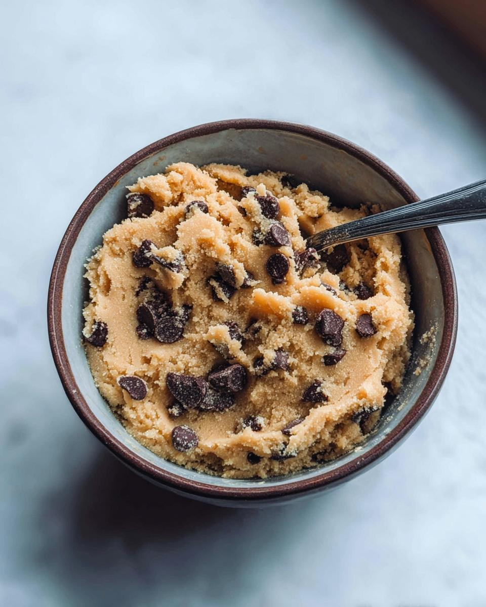 A bowl of Delicious Protein Cookie Dough loaded with chocolate chips, with a spoon in the bowl.