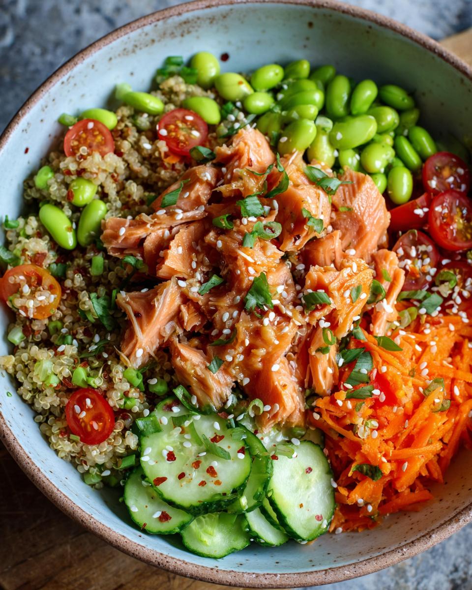Close-up of a healthy salmon power bowl featuring flaked salmon, quinoa, edamame, shredded carrots, cucumber slices, and cherry tomatoes.