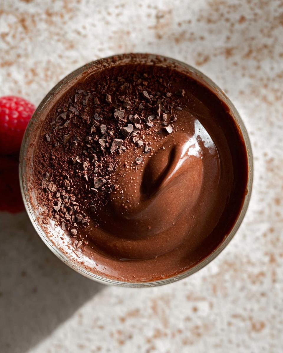 A close-up overhead view of a glass filled with rich, dark High Protein Chocolate Pudding, topped with cacao nibs and a raspberry on the side.