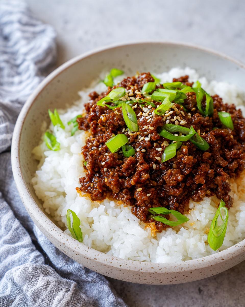 A close-up of a Korean beef rice bowl with seasoned ground beef over white rice, topped with green onions and sesame seeds.